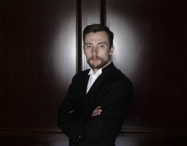 A man in a suit stands facing forward with his arms crossed, a spotlight shines on him, the background is dark
