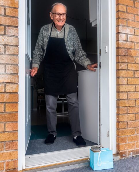 Elderly man standing in his doorway, with a package of food on his doorstep